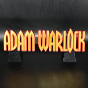 May include: A decorative sign with the text "ADAM WARLOCK" in a bold, stylized font. The letters are yellow with a red outline, set against a black background. The sign is supported by two small, black stands.