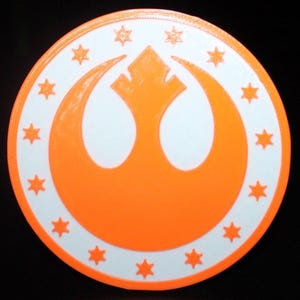 May include: A round, orange and white emblem featuring a stylized bird-like symbol at the center, surrounded by a ring of stars. The emblem is set against a black background. The design is reminiscent of a popular science fiction franchise.