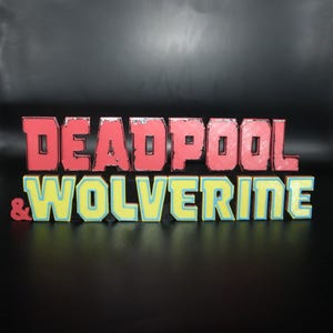 Peut inclure: Panneau 3D coloré avec les mots "DEADPOOL & WOLVERINE". Le texte rouge est au-dessus du texte jaune et bleu. L'esperluette est rouge. Le tout est sur un fond sombre.