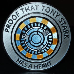 Könnte beinhalten: Ein silberfarbenes, kreisförmiges Emblem mit dem Text "PROOF THAT TONY STARK HAS A HEART". Das Zentrum zeigt ein blaues und goldenes Design, das einem Reaktorkern ähnelt. Das Emblem ist wahrscheinlich ein Sammler- oder Fanartikel.