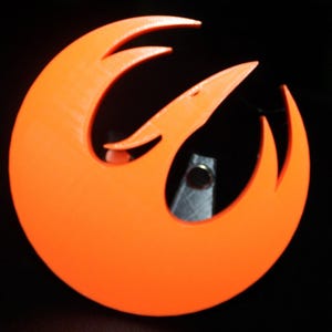 Könnte beinhalten: Hellorangefarbenes Emblem mit einem stilisierten Vogeldesign, vor schwarzem Hintergrund. Das Emblem hat eine kreisförmige Form mit gebogenen, spitzen Elementen. Das Design erinnert an einen Phönix oder einen stilisierten Vogel im Flug.