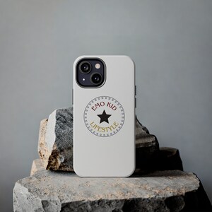 May include: A white phone case featuring a black camera module and the text "EMO KID LIFESTYLE" in a circular design with a black star. The case is displayed on a textured gray stone surface.