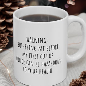 Può includere: Tazza da caffè in ceramica bianca riempita di liquido scuro, con il testo: "WARNING: BOTHERING ME BEFORE MY FIRST CUP OF COFFEE CAN BE HAZARDOUS TO YOUR HEALTH." La tazza è su una superficie grigio chiaro.