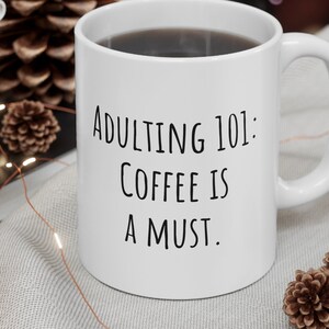 Puede incluir: Taza de café de cerámica blanca con el texto "Adulting 101: Coffee is a Must." La taza está llena de café oscuro y tiene un asa. La taza está sobre una superficie gris claro con elementos decorativos.