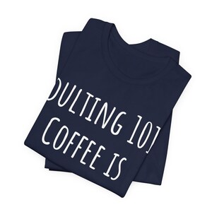 May include: Navy blue t-shirt with the text "Adulting 101 Coffee Is" in white, stacked vertically. The shirt is folded, showing the collar and part of the front. The fabric appears soft and comfortable.