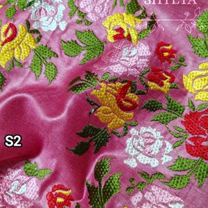 Może przedstawiać: Różowy materiał z haftowanym wzorem kwiatowym w kolorze żółtym, czerwonym i białym. Materiał ma subtelny połysk i miękką fakturę. Tekst "SHYETA" jest wyszyty na materiale na biało.