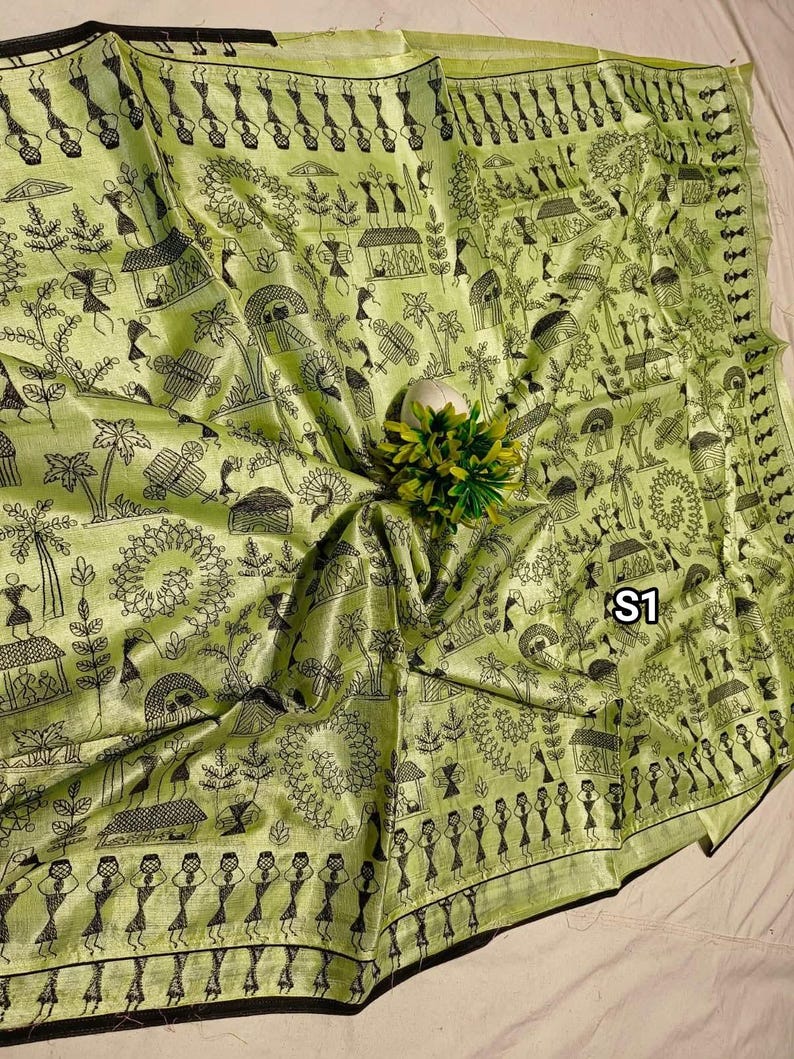 May include: A light green textile with a black illustrated village scene. The fabric has a shiny appearance and features detailed drawings of people, houses, and trees. The edge of the textile is trimmed with a black border. The text "S1" is visible.