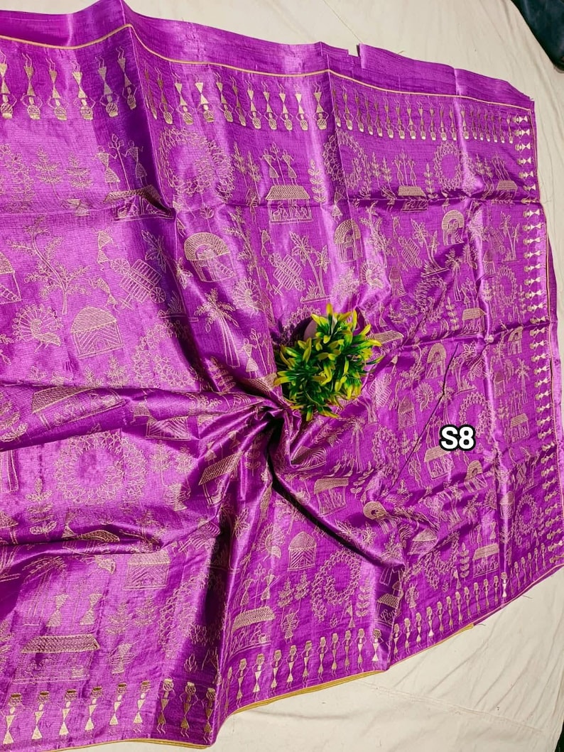 May include: A vibrant purple silk saree with intricate gold-colored patterns. The saree features detailed designs along the borders and across the fabric, with a small green plant in the center. The tag "S8" is visible.