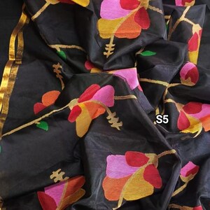 May include: Black sheer saree with gold trim and colorful floral designs. The saree has a textured, almost quilted look. The saree is folded and draped over a surface.