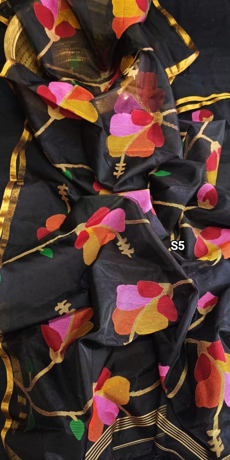 May include: Black sheer saree with gold trim and colorful floral designs. The saree has a textured, almost quilted look. The saree is folded and draped over a surface.