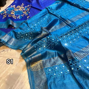 May include: A blue and purple silk saree with gold embroidery and mirror work. The saree features a geometric pattern and is decorated with small, round mirrors. The saree is folded and displayed on a wooden surface. S1 is written on the surface.