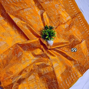 May include: Mustard yellow silk textile with intricate white embroidered patterns. The textile features detailed scenes and designs, with the text "S9" visible in the corner. A small potted plant sits on top of the textile.