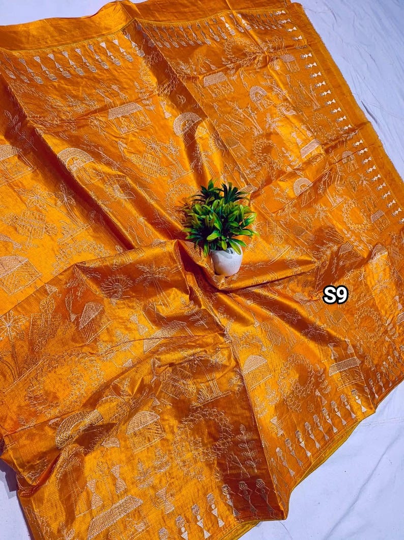 May include: Mustard yellow silk textile with intricate white embroidered patterns. The textile features detailed scenes and designs, with the text "S9" visible in the corner. A small potted plant sits on top of the textile.