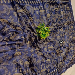 May include: A navy blue textile with a gold-colored pattern featuring stylized figures, trees, and huts. The fabric has a subtle sheen and a decorative border. The text "S2" is visible on the fabric.