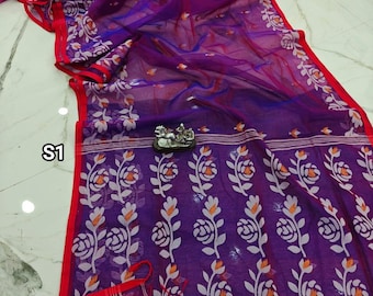 May include: A sheer purple saree with a floral pattern in white and orange. The saree has a red border and is draped on a white surface. The design includes a small decorative figurine.