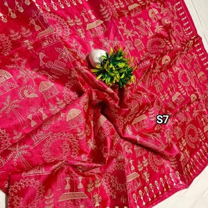 May include: A vibrant pink textile with intricate gold embroidery depicting village scenes and figures. The fabric is draped, showcasing the detailed patterns and textures. The design includes houses, trees, and people, creating a narrative feel.