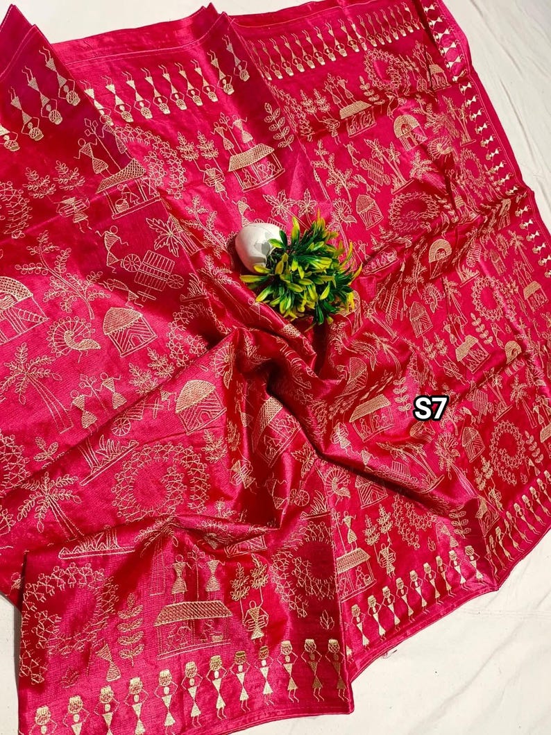 May include: A vibrant pink textile with intricate gold embroidery depicting village scenes and figures. The fabric is draped, showcasing the detailed patterns and textures. The design includes houses, trees, and people, creating a narrative feel.