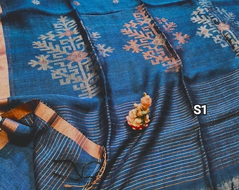 May include: A blue linen saree with a geometric pattern and a gold border. The saree has a fringe at the bottom and the text "S1" is visible in the image.