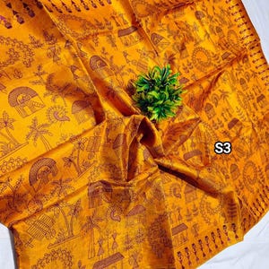 May include: Mustard yellow textile with a detailed brown pattern featuring village scenes, trees, and figures. The fabric is folded, with a small green plant in the center. The text "S3" is visible in the corner.