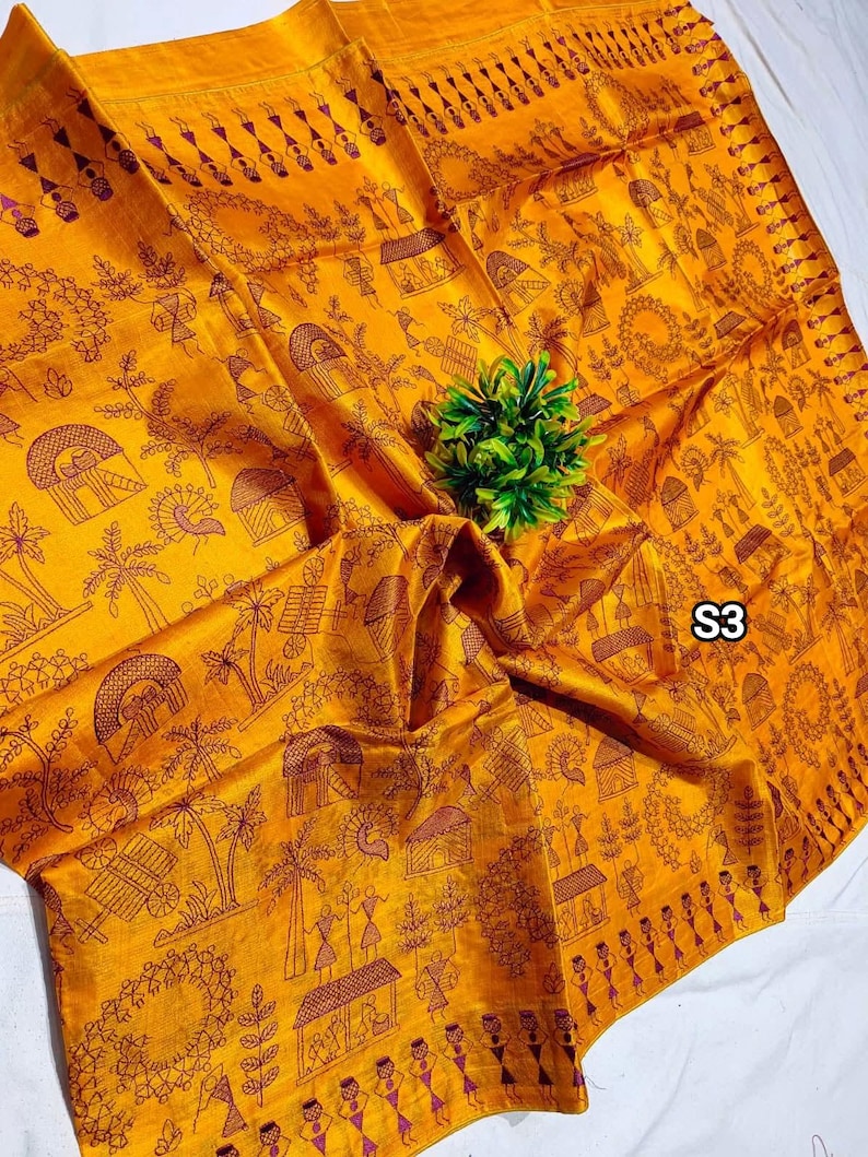 May include: Mustard yellow textile with a detailed brown pattern featuring village scenes, trees, and figures. The fabric is folded, with a small green plant in the center. The text "S3" is visible in the corner.