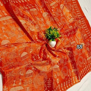 May include: An orange textile with a detailed gold pattern featuring village scenes, figures, and geometric shapes. The fabric is draped, with a small white vase holding a green plant in the center. The text "S5" is visible.