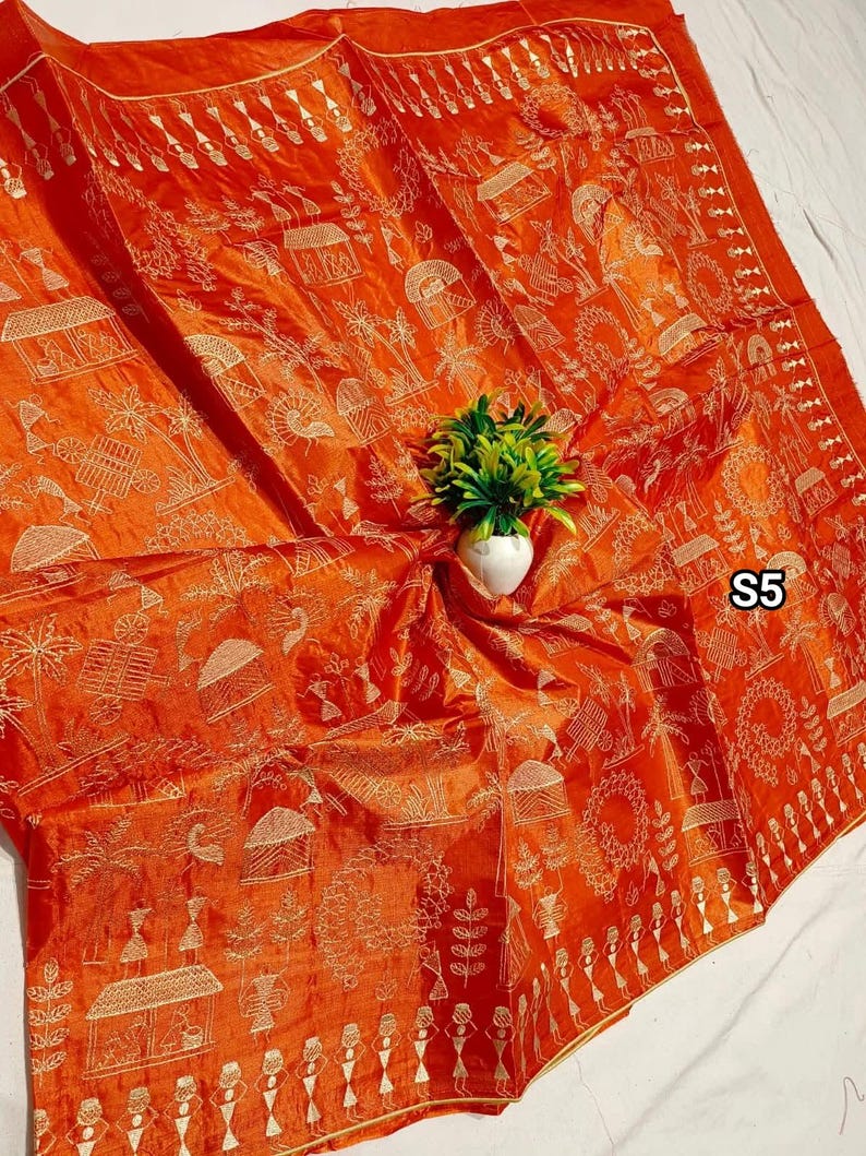 May include: An orange textile with a detailed gold pattern featuring village scenes, figures, and geometric shapes. The fabric is draped, with a small white vase holding a green plant in the center. The text "S5" is visible.