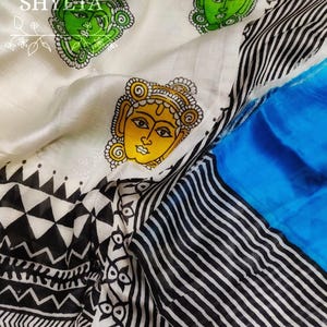 May include: White silk saree with black and white geometric patterns and blue trim. The saree features hand-painted faces in green, yellow, and black.