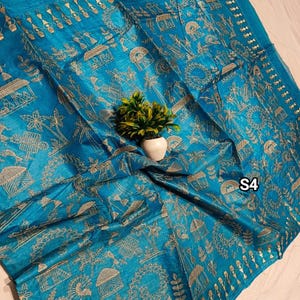 May include: A vibrant turquoise textile with intricate gold-colored embroidery. The design features stylized houses, trees, and figures, creating a detailed pattern. The textile is draped, and a small plant in a white pot is placed on top. The text "S4" is visible.