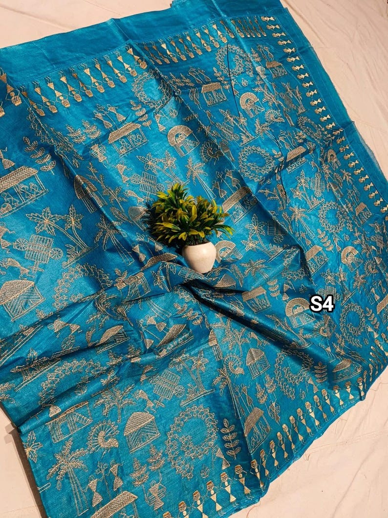 May include: A vibrant turquoise textile with intricate gold-colored embroidery. The design features stylized houses, trees, and figures, creating a detailed pattern. The textile is draped, and a small plant in a white pot is placed on top. The text "S4" is visible.