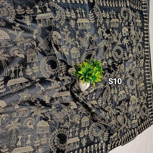 May include: Black fabric with intricate gold embroidery depicting village scenes, figures, and geometric patterns. The textile features a detailed border and the text "S10" in the upper right corner. A small green plant in a white pot is in the center.