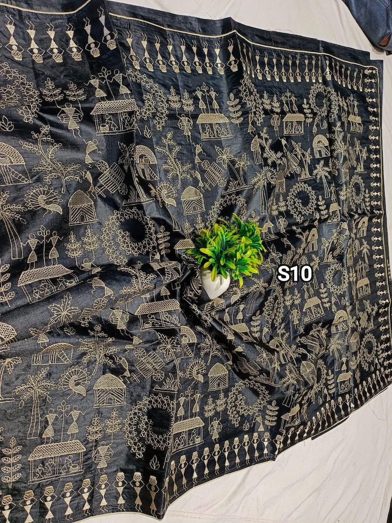 May include: Black fabric with intricate gold embroidery depicting village scenes, figures, and geometric patterns. The textile features a detailed border and the text "S10" in the upper right corner. A small green plant in a white pot is in the center.