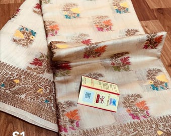 May include: Cream-colored textile with floral patterns in orange, pink, blue, and yellow. The fabric has a gold border with intricate designs. A small box with text is visible. The textile is folded and displayed on a wooden surface.