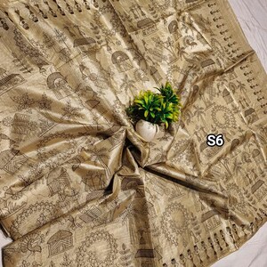May include: A gold-colored textile with a detailed black line drawing of a village scene. The fabric is draped, with a small white vase holding green plants in the center. The text "S6" is visible.