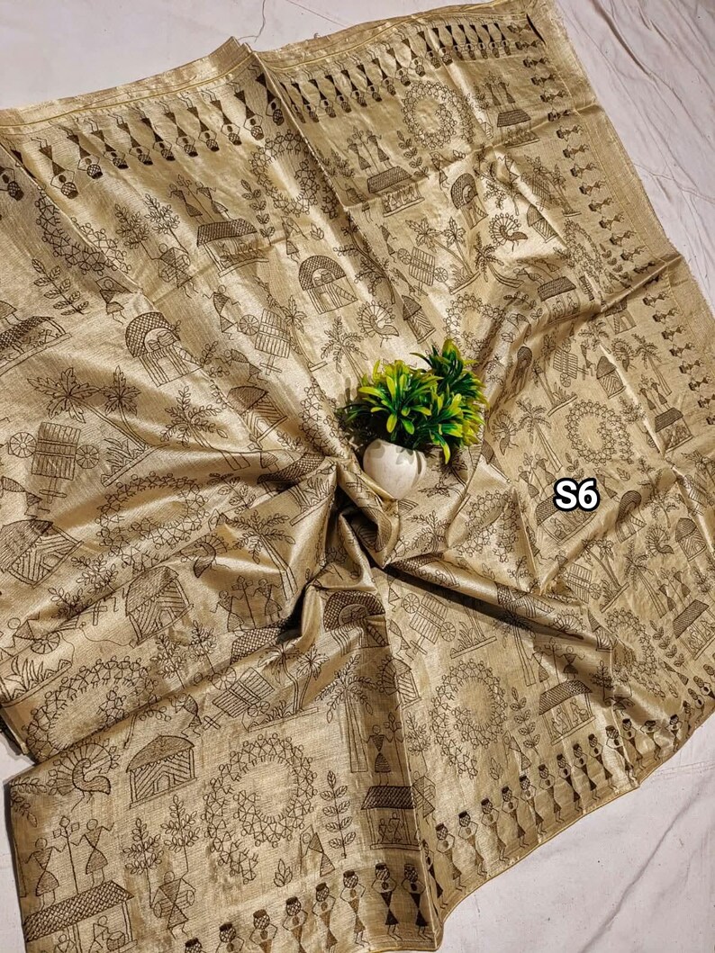 May include: A gold-colored textile with a detailed black line drawing of a village scene. The fabric is draped, with a small white vase holding green plants in the center. The text "S6" is visible.