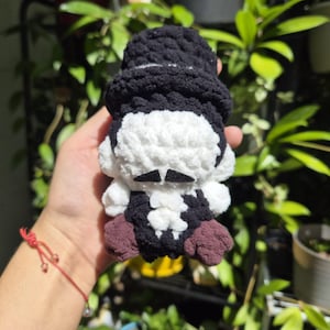 May include: A small, handmade crochet plush doll featuring a black top hat, a white head with a black felt mustache, a black vest over a white shirt, and brown feet. The figure is crafted from soft, chunky yarn with a textured, looped appearance.