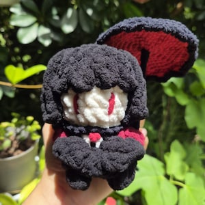 May include: A handmade crochet plush doll featuring a black textured yarn body, a white face with red embroidered eyes, and a small red accent at the center. The doll has a large, circular black and red mushroom-like cap attached to the top.