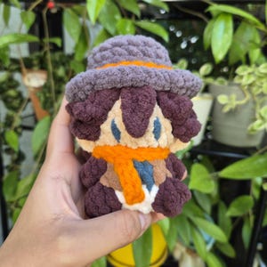 May include: A small, handmade crochet plush doll featuring a dark purple hat with an orange band, dark brown hair, a beige face with blue eyes, and a bright orange scarf wrapped around its neck. The figure has a soft, textured yarn construction.