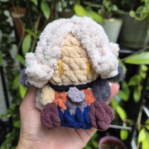 May include: A small, handmade crochet plush toy featuring a character with voluminous, textured cream-colored yarn hair, a tan face with yellow embroidered eyes, and a multi-colored body made of dark blue, maroon, orange, and grey yarn segments.