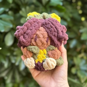May include: A handmade, textured crochet item in a variety of colors, including purple, yellow, green, and beige. The item has a rounded shape and appears to be a decorative piece or small sculpture. The soft, plush texture is visible.