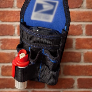 May include: Black and blue utility belt with multiple pockets and a red and white item in the front pocket. The belt has a blue top with a white logo. The belt is hanging on a brick wall.