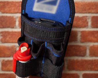 May include: Black and blue utility belt with multiple pockets and a red and white item in the front pocket. The belt has a blue top with a white logo. The belt is hanging on a brick wall.