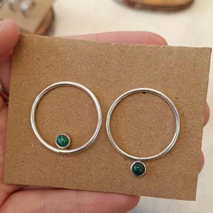 Può includere: Un paio di orecchini a cerchio in argento, ciascuno con una piccola pietra rotonda verde scuro. Gli orecchini sono esposti su un cartoncino marrone. I cerchi hanno un diametro di circa 2,5 cm.