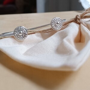 Op de afbeelding: Een zilveren armband met twee decoratieve ronde accenten. De armband ligt op een wit stoffen oppervlak.
