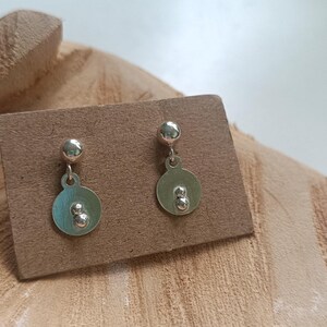 Op de afbeelding: Een paar zilverkleurige oorbellen met een kleine bolvormige knop bevestigd aan een platte ronde schijf. Elke schijf is versierd met twee kleine verhoogde bolvormige accenten die verticaal in het midden zijn geplaatst voor een glanzend metalen ontwerp.
