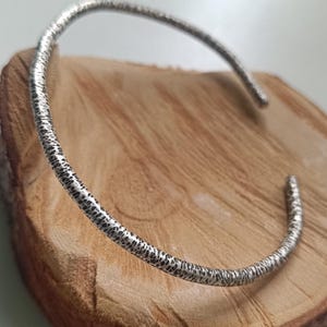 Peut inclure: Un bracelet manchette argenté avec une surface texturée. Le bracelet ouvert est présenté sur une tranche de bois, mettant en valeur son design martelé. Le bijou est un accessoire simple mais élégant.