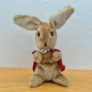 May include: A plush rabbit toy with light brown fur, long upright ears, and dark plastic eyes. The rabbit is wearing a small red cape with a white trim pattern, secured around the neck with a white ribbon. The toy is in a seated position.