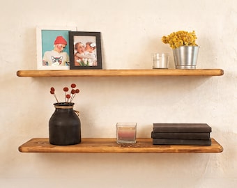 May include: Two wooden floating shelves with various decorative items. The top shelf holds framed photos, a small glass, and a metal bucket with yellow flowers. The bottom shelf displays a black vase, a candle, and books.