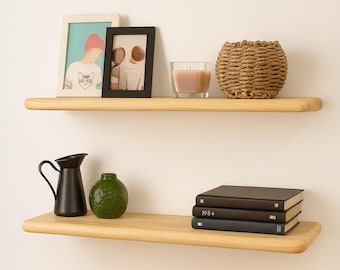 May include: Two light-colored wooden floating shelves mounted on a white wall. The top shelf holds framed photos, a candle, and a woven basket. The bottom shelf displays a black pitcher, a green vase, and stacked books.