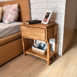 May include: A wooden nightstand with a drawer and a lower shelf. The nightstand is made of light-colored wood and has rounded edges. A book, glasses, and a framed photo sit on top. A basket with toiletries is on the shelf below. The nightstand is next to a bed.