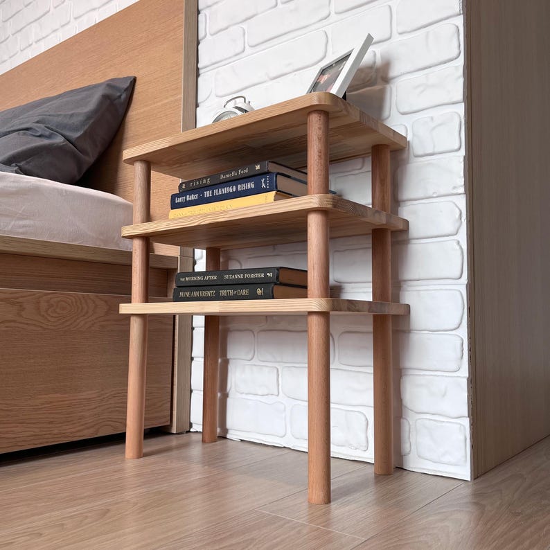 Puede incluir: Una mesita de noche de madera de tres niveles con acabado natural. La mesa tiene una parte superior y estantes rectangulares sostenidos por patas cil&iacute;ndricas. Libros y una foto enmarcada se exhiben en los estantes. La mesa est&aacute; junto a una pared de ladrillos blancos.